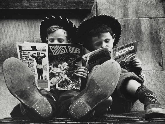 Two children wearing cowboy hats and boots, reading comics.