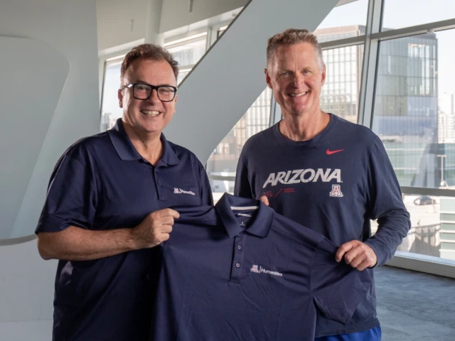 Two men smiling and holding up a blue polo that has a Block A and says "Humanities"
