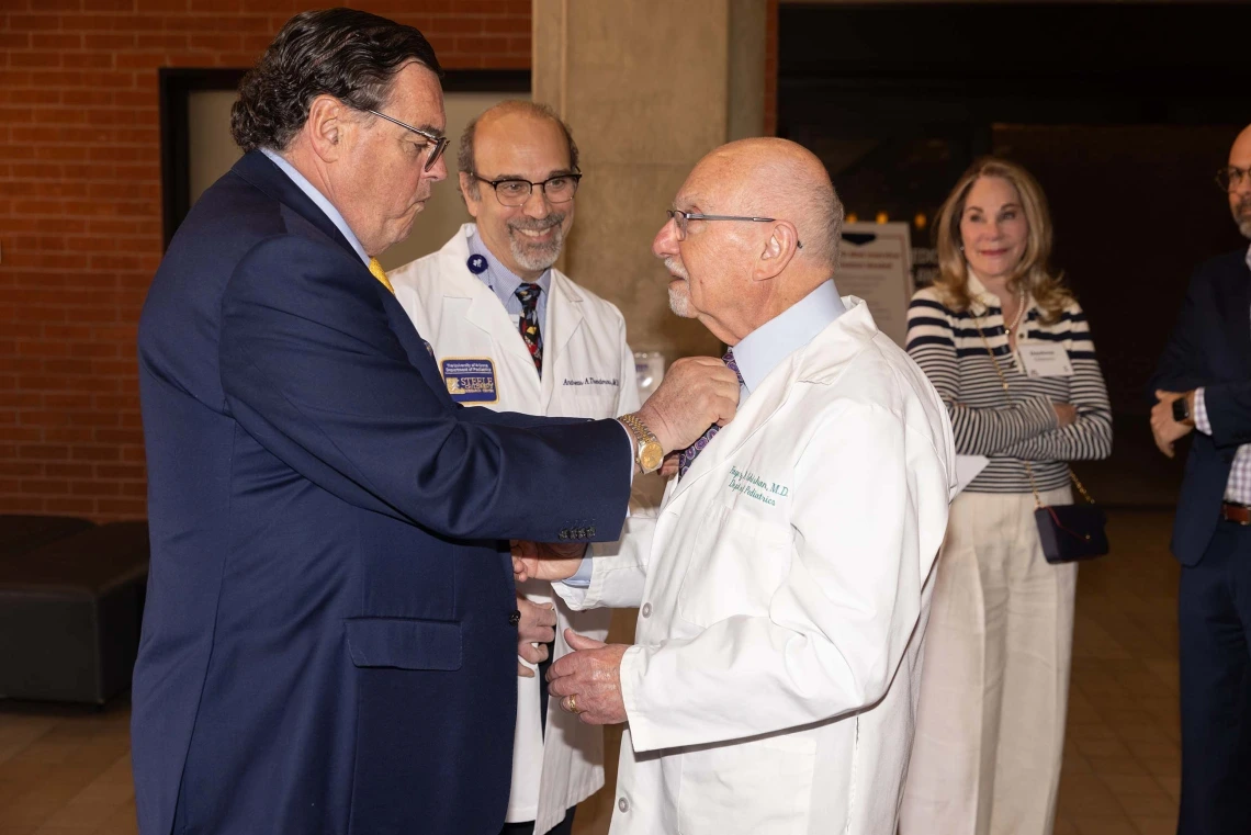 A man adjusting the tie of another, wearing a white coat, while three people smile 