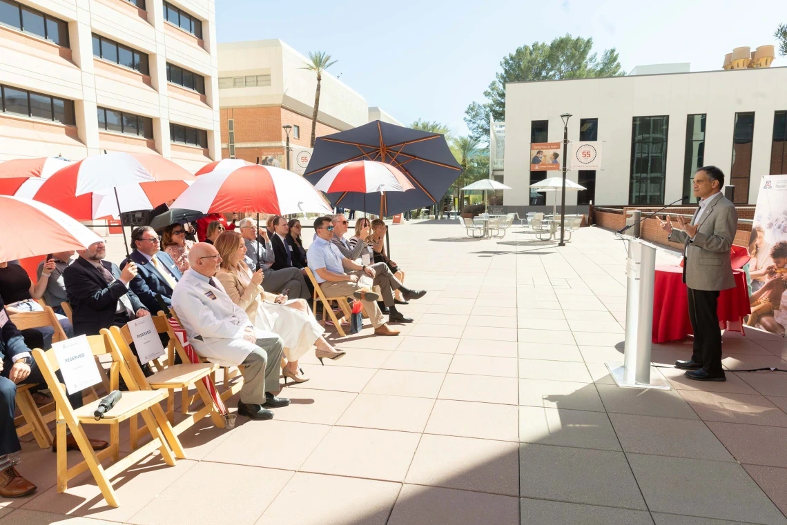People sitting outside under an umbrella while President Garimella speaks.
