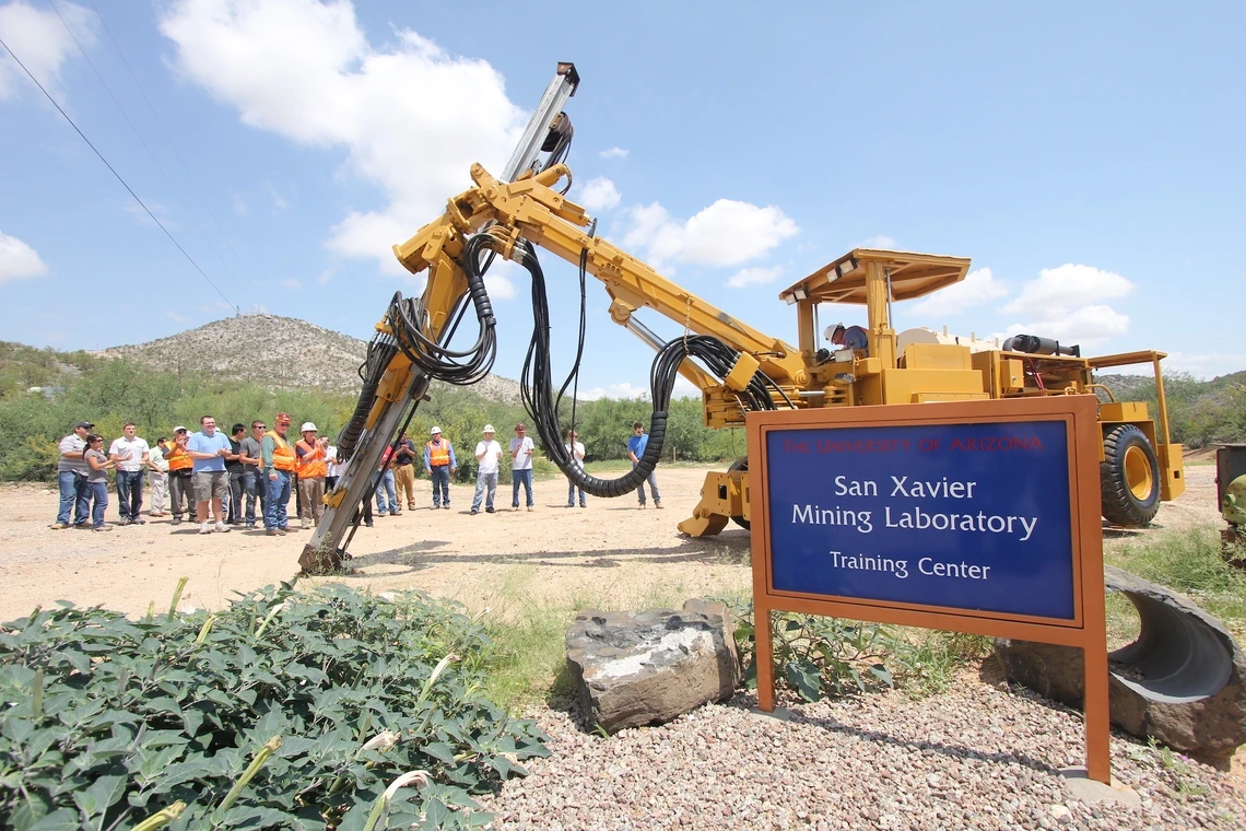 A group of people looking at an excavator behind a sign that says, "San Xavier Mining Laboratory."