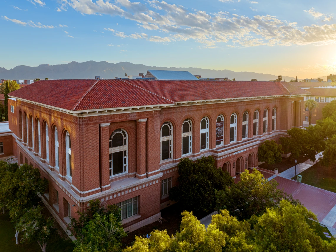 An image of a building on the University of Arizona campus with the iconic red brick. The sun is seen setting in the background on the right side of the image.