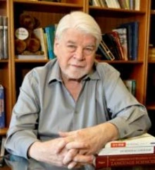 A man smiling with his hands crossed, in front of a book shelf